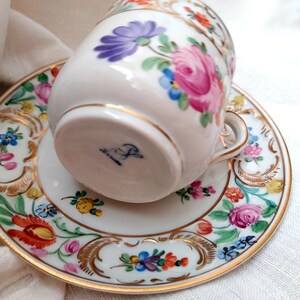 May include: A white porcelain teacup and saucer with a floral pattern in shades of pink, blue, yellow, and orange. The cup has a gold rim and the saucer has a gold rim and a gold floral design. The bottom of the cup has a blue and white mark.
