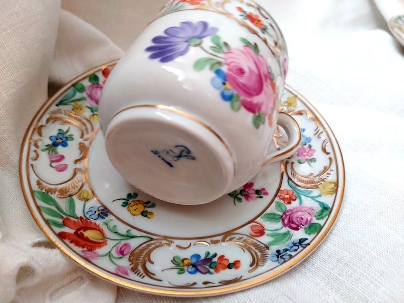May include: A white porcelain teacup and saucer with a floral pattern in shades of pink, blue, yellow, and orange. The cup has a gold rim and the saucer has a gold rim and a gold floral design. The bottom of the cup has a blue and white mark.