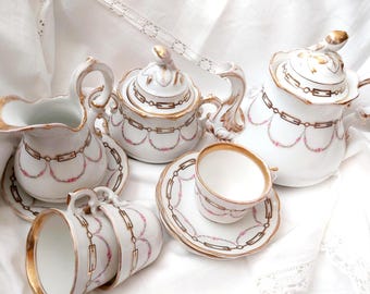 May include: A white porcelain tea set featuring a delicate gold-trimmed chain pattern and small pink floral swags. The set includes a teapot, a lidded sugar bowl, a creamer, and two teacups with matching saucers, all with ornate gold-painted handles.