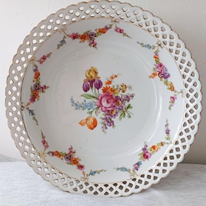 Puede incluir: Bol de porcelana blanca para servir con un delicado borde calado y un diseño floral. El bol presenta un ramo central de flores coloridas, incluyendo rosas, iris y tulipanes. El borde está adornado con detalles dorados.