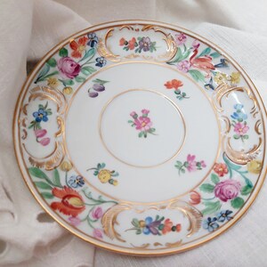 May include: A white porcelain saucer with a gold rim and a floral design. The flowers are painted in shades of pink, red, yellow, blue, and green.