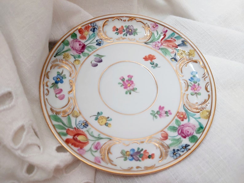 May include: A white porcelain saucer with a gold rim and a floral design. The flowers are painted in shades of pink, red, yellow, blue, and green.