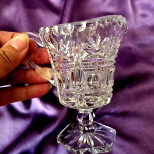 May include: A clear pressed glass creamer featuring a geometric pattern of starbursts and vertical lines. The vessel has a scalloped rim, a curved handle, and a hexagonal base with a starburst design. The glass is transparent and reflective.
