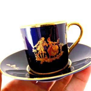 May include: A dark blue teacup and saucer set with gold trim. The teacup and saucer feature a gold illustration of a couple in period clothing. The teacup handle and rim are also gold.