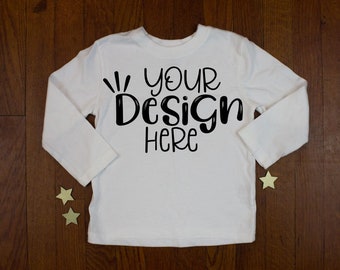 Puede incluir: Camiseta blanca de manga larga con el texto "YOUR Design HERE" en escritura negra. La prenda se presenta sobre una superficie de madera oscura, con tres pequeñas decoraciones en forma de estrella. Una camiseta en blanco para diseños personalizados.