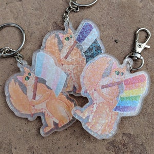 Puede incluir: Tres llaveros de gato naranja con detalles de purpurina. Cada gato sostiene una bandera del orgullo diferente: una bandera transgénero, una bandera bisexual y una bandera arcoíris.