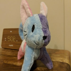 K&ouml;nnte beinhalten: Ein Pl&uuml;schtier in Form einer Katze mit blauem und violettem K&ouml;rper, rosa Ohren und schwarzen Augen. Das Spielzeug ist aus weichem, flauschigem Stoff.