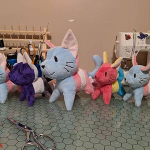 K&ouml;nnte beinhalten: F&uuml;nf Pl&uuml;schkatzenspielzeuge mit unterschiedlich farbigem Fell und Streifen. Die Katzen sind in einer Reihe auf einem Tisch mit einem sechseckigen Muster angeordnet.