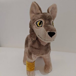 May include: A plush wolf toy in shades of brown and tan. The toy features large yellow eyes, a black nose, and a yellow band around one leg. The soft toy is made from a plush material.