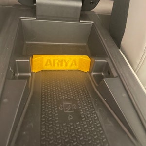 May include: A yellow plastic nameplate with the word "ARIYA" printed on it. The nameplate is sitting in a black plastic car console.