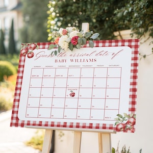May include: A wooden easel displays a calendar with a red and white gingham border, adorned with floral accents and apples. The calendar, with the text "Guess the arrival date of BABY WILLIAMS," is set against a blurred outdoor backdrop.