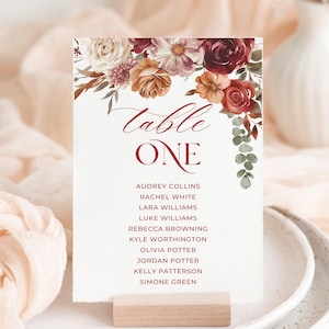 May include: A wedding table card with floral accents. The card reads "Table One" in burgundy script, with a list of names below. The top of the card is decorated with flowers in shades of burgundy, cream, and orange. The card is held by a small wooden block.
