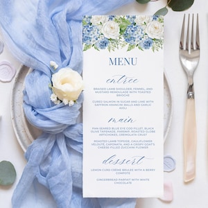 May include: A wedding menu card with a floral design of blue hydrangeas and white roses. The menu card is on a white surface with a light blue fabric, silverware, and a ring box.