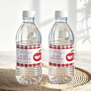May include: Two clear water bottles with white caps. Each bottle has a label with a red and white gingham pattern, the text "Audrey's BABY SHOWER", and a red apple with a heart cutout. The bottles are on a woven mat.