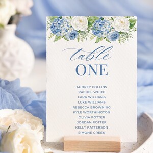 May include: A white table card with blue floral accents, displaying the text "Table ONE" and guest names. The card is held by a small wooden block. The background features white flowers and blue fabric.