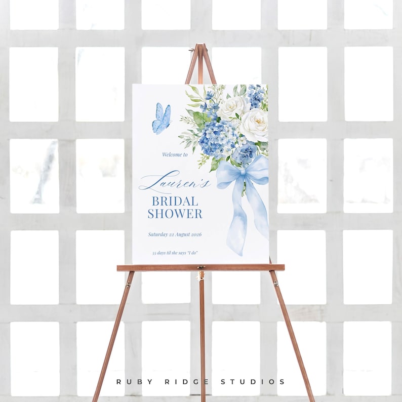 K&ouml;nnte beinhalten: Ein wei&szlig;es Schild auf einer Holzstaffelei mit der Aufschrift "Lauren's Bridal Shower". Das Schild zeigt ein Aquarelldesign mit blauen Blumen, einem Schmetterling und einer Schleife. Das Datum der Party ist der 22. August 2026.