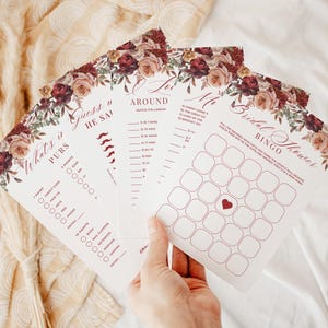 Puede incluir: Un conjunto de tarjetas de juegos para despedida de soltera con detalles florales. Las tarjetas presentan varios juegos, incluyendo "What's in Purses" y "Bridal Shower Bingo". El diseño incorpora rosas burdeos y rosadas.