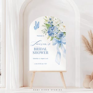 K&ouml;nnte beinhalten: Ein wei&szlig;es, bogenf&ouml;rmiges Schild auf einer Holzstaffelei mit einem Blumendesign aus blauen Hortensien, wei&szlig;en Rosen und einer blauen Schleife. Auf dem Schild steht "Willkommen zu Laurens Bridal Shower" mit Datum und Countdown. Ein blauer Schmetterling ist ebenfalls abgebildet.