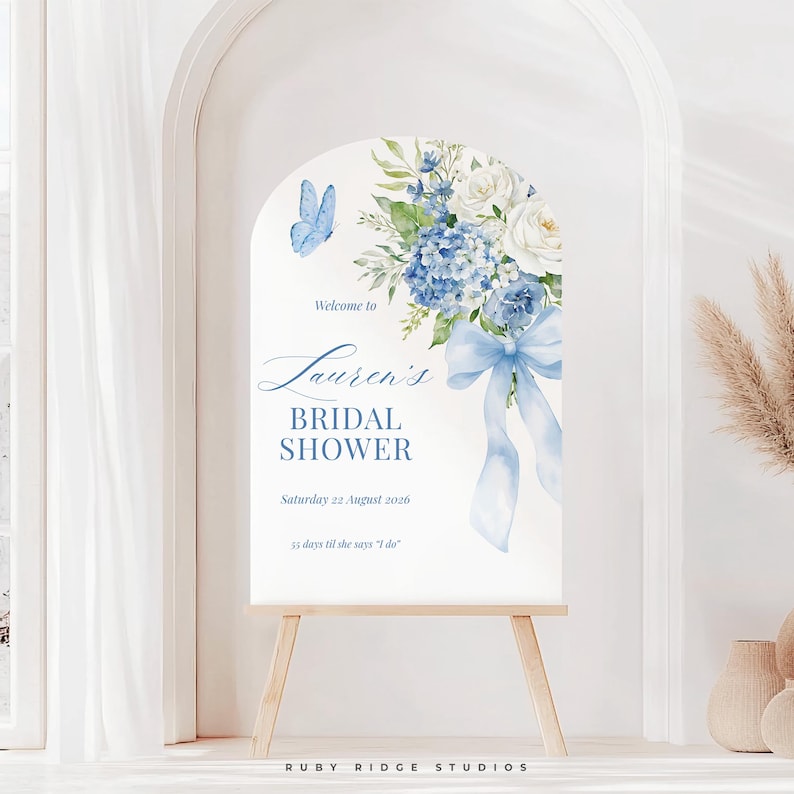 K&ouml;nnte beinhalten: Ein wei&szlig;es, bogenf&ouml;rmiges Schild auf einer Holzstaffelei mit einem Blumendesign aus blauen Hortensien, wei&szlig;en Rosen und einer blauen Schleife. Auf dem Schild steht "Willkommen zu Laurens Bridal Shower" mit Datum und Countdown. Ein blauer Schmetterling ist ebenfalls abgebildet.