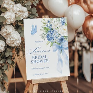 K&ouml;nnte beinhalten: Ein wei&szlig;es Schild auf einer Holzstaffelei mit einem Blumendesign aus blauen Hortensien, wei&szlig;en Rosen und einem blauen Schmetterling. Auf dem Schild steht "Willkommen zu Laurens Bridal Shower" mit dem Datum "Samstag, 22. August 2026" und "55 Tage bis sie 'Ja' sagt".