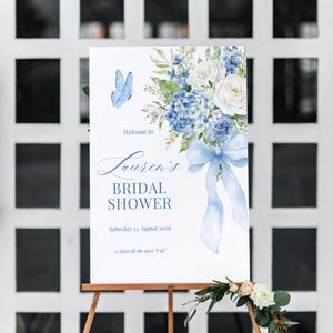 K&ouml;nnte beinhalten: Ein Schild auf einem Holzstaffelei, das zu einer Brautparty einl&auml;dt. Es zeigt blaue Blumenillustrationen, einen Schmetterling und den Text "Welcome to Lauren's Bridal Shower, Samstag 22. August 2026, 55 Tage bis sie 'Ja' sagt".