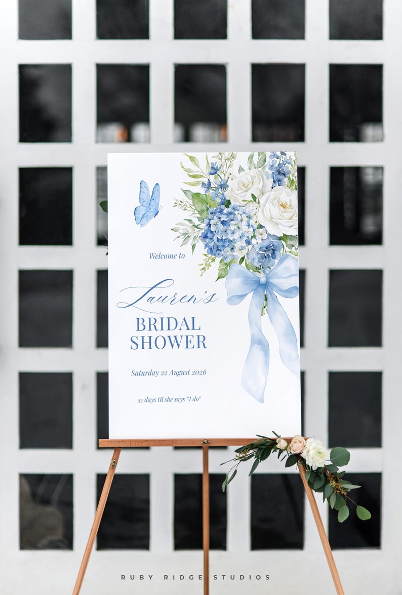K&ouml;nnte beinhalten: Ein Schild auf einem Holzstaffelei, das zu einer Brautparty einl&auml;dt. Es zeigt blaue Blumenillustrationen, einen Schmetterling und den Text "Welcome to Lauren's Bridal Shower, Samstag 22. August 2026, 55 Tage bis sie 'Ja' sagt".