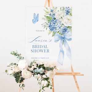 K&ouml;nnte beinhalten: Ein Schild f&uuml;r eine Brautparty auf einer h&ouml;lzernen Staffelei. Das Schild zeigt ein Aquarelldesign mit blauen Blumen, einem Schmetterling und dem Text "Lauren's Bridal Shower". Eine Blumengesteck mit wei&szlig;en Blumen und Gr&uuml;n befindet sich am Fu&szlig;.