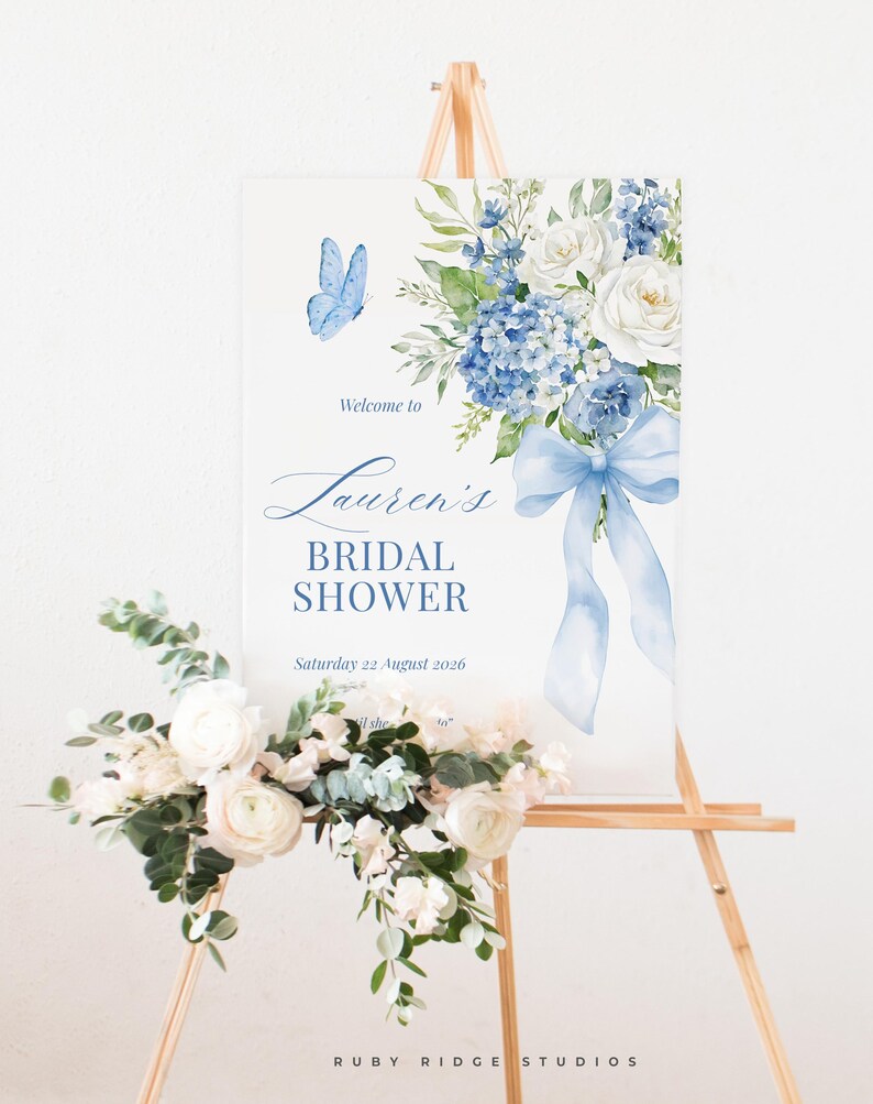 K&ouml;nnte beinhalten: Ein Schild f&uuml;r eine Brautparty auf einer h&ouml;lzernen Staffelei. Das Schild zeigt ein Aquarelldesign mit blauen Blumen, einem Schmetterling und dem Text "Lauren's Bridal Shower". Eine Blumengesteck mit wei&szlig;en Blumen und Gr&uuml;n befindet sich am Fu&szlig;.