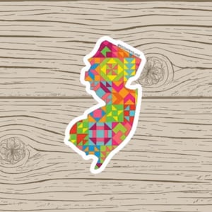 May include: A sticker in the shape of New Jersey, filled with a colorful geometric pattern of triangles and squares in shades of red, orange, yellow, green, blue, and pink. The sticker is on a wooden background.
