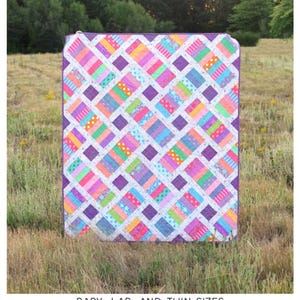 May include: A colorful quilt titled "The Iris Quilt" by Erica Jackman, displayed outdoors. The quilt features a patchwork design with squares and rectangles in various colors like pink, purple, blue, green, and yellow. The text "Baby, Lap, and Twin Sizes" is visible.