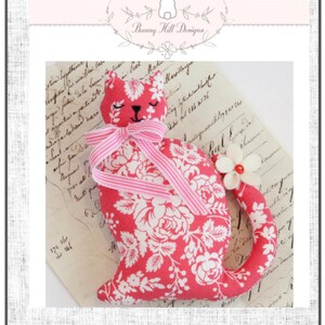 May include: A handmade cat-shaped plush toy in red fabric with a white floral pattern. It features a pink striped bow, a white flower on the tail, and the text "Blossom Pattern". The Bunny Hill Designs logo is at the top.