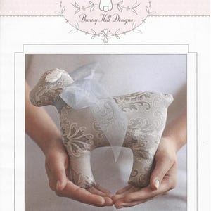 May include: A stuffed lamb toy with a damask pattern in shades of gray and white, adorned with a light blue ribbon. The toy is held in cupped hands. The text "Lollipop" is at the bottom of the image, and the Bunny Hill Designs logo is at the top.