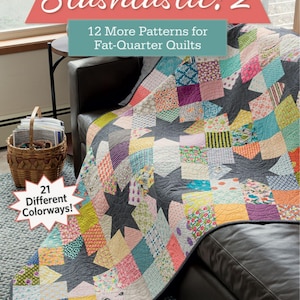 May include: A colorful quilt with star patterns and patchwork squares is draped over a dark leather couch. The quilt features various fabric patterns and colors. The book "Stashtastic! 2" is visible, with text indicating 12 quilt patterns and 21 colorways.
