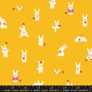 May include: A mustard yellow fabric featuring a repeating pattern of white cartoon rabbits in various poses, some holding small red tomatoes. A black ruler at the bottom shows measurements in inches, with the text Ruby Star Society printed repeatedly.