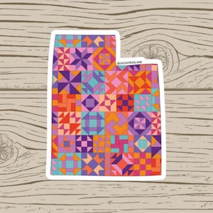 May include: A sticker in the shape of Utah, featuring a colorful patchwork quilt design. The quilt pattern incorporates geometric shapes in shades of orange, purple, pink, and teal, set against a white border. The sticker is on a wood grain background.