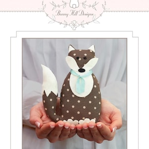 May include: A handmade stuffed fox doll with a brown polka-dot body, white accents, and a light blue scarf. The doll is held in open hands and features the text "Foxly" at the bottom. The Bunny Hill Designs logo is at the top.