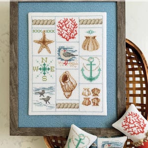 May include: A framed cross-stitch sampler titled "Shoreline Sampler" featuring nautical motifs like a starfish, coral, seashell, seagull, anchor, and compass. The sampler is displayed with decorative pillows in a basket, all with similar coastal designs.