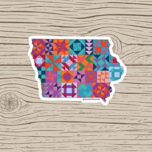 May include: A sticker in the shape of Iowa, featuring a colorful patchwork design. The design incorporates various geometric shapes in shades of orange, pink, blue, and purple. The text "@SASSAFRASLANE" is at the bottom.
