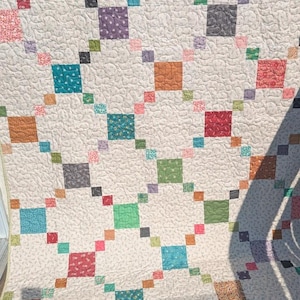 May include: A patchwork quilt featuring a cream-colored background with a swirling, continuous line quilting pattern. The design consists of a grid of solid and patterned squares in various colors including teal, orange, red, green, and gray.