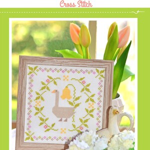 Peut inclure: Un modèle de point de croix encadré intitulé Gertrude The Goose Cross Stitch représentant une oie centrale entourée d'une couronne florale de feuilles vertes, de fleurs roses et d'une fleur jaune en forme de cloche sur tissu blanc.