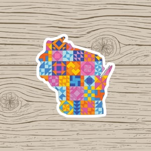 May include: A sticker in the shape of Wisconsin, filled with a colorful patchwork quilt design. The quilt features various geometric patterns in shades of blue, pink, orange, and yellow. The sticker is on a wooden background.