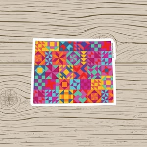 May include: A colorful sticker featuring a patchwork quilt design. The sticker showcases a vibrant array of geometric shapes in shades of red, orange, yellow, blue, and purple. The sticker is rectangular with a white border and is placed on a wooden surface.