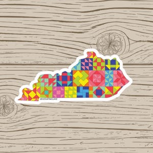 May include: A sticker in the shape of the state of Kentucky, filled with a colorful patchwork quilt design. The sticker has a white border and the text "@SASSAFRASLANE" is printed on the bottom.