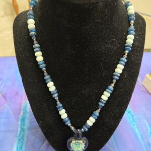 May include: A beaded necklace with a heart-shaped pendant. The necklace features blue, white, and clear beads. The pendant is a dark blue glass heart with a light green and white floral design inside. The necklace is displayed on a black bust.