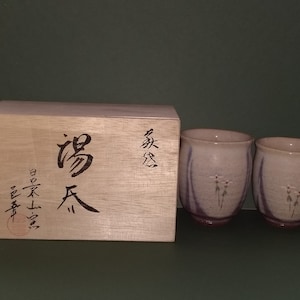 May include: Two small, pale pink ceramic teacups with purple and white floral designs, sitting on a wooden box with Japanese characters. The box is labeled "高天" and "款" in black ink.