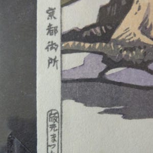 Pu&ograve; includere: Un dettaglio di una stampa xilografica giapponese con testo verticale nero su una striscia bianca. La stampa presenta un paesaggio con toni tenui di beige, grigio e giallo. Il testo recita "京都御所" e un timbro circolare con caratteri giapponesi.