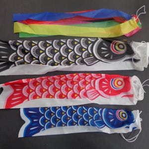 May include: A set of four carp-shaped wind socks, known as koinobori, featuring one with multi-colored streamers and three with fish-scale patterns in black, red, and blue. Each fish has a circular eye design and a white string loop at the mouth.