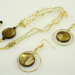 May include: A gold necklace with a brown and white shell pendant and matching earrings. The necklace is made of a chain with small clear and gold beads. The earrings are hoop style with a brown and white shell pendant.