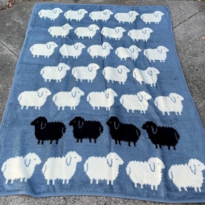 May include: A blue bath towel featuring a pattern of white and black sheep. The towel is rectangular and has a soft, plush texture. The sheep are evenly spaced across the surface.