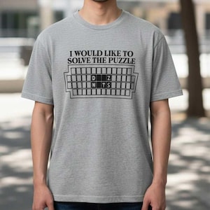 May include: A grey t-shirt featuring the text "I WOULD LIKE TO SOLVE THE PUZZLE" above a game board design in black. The board shows the letters D, Z, N, and TS. The shirt has short sleeves.